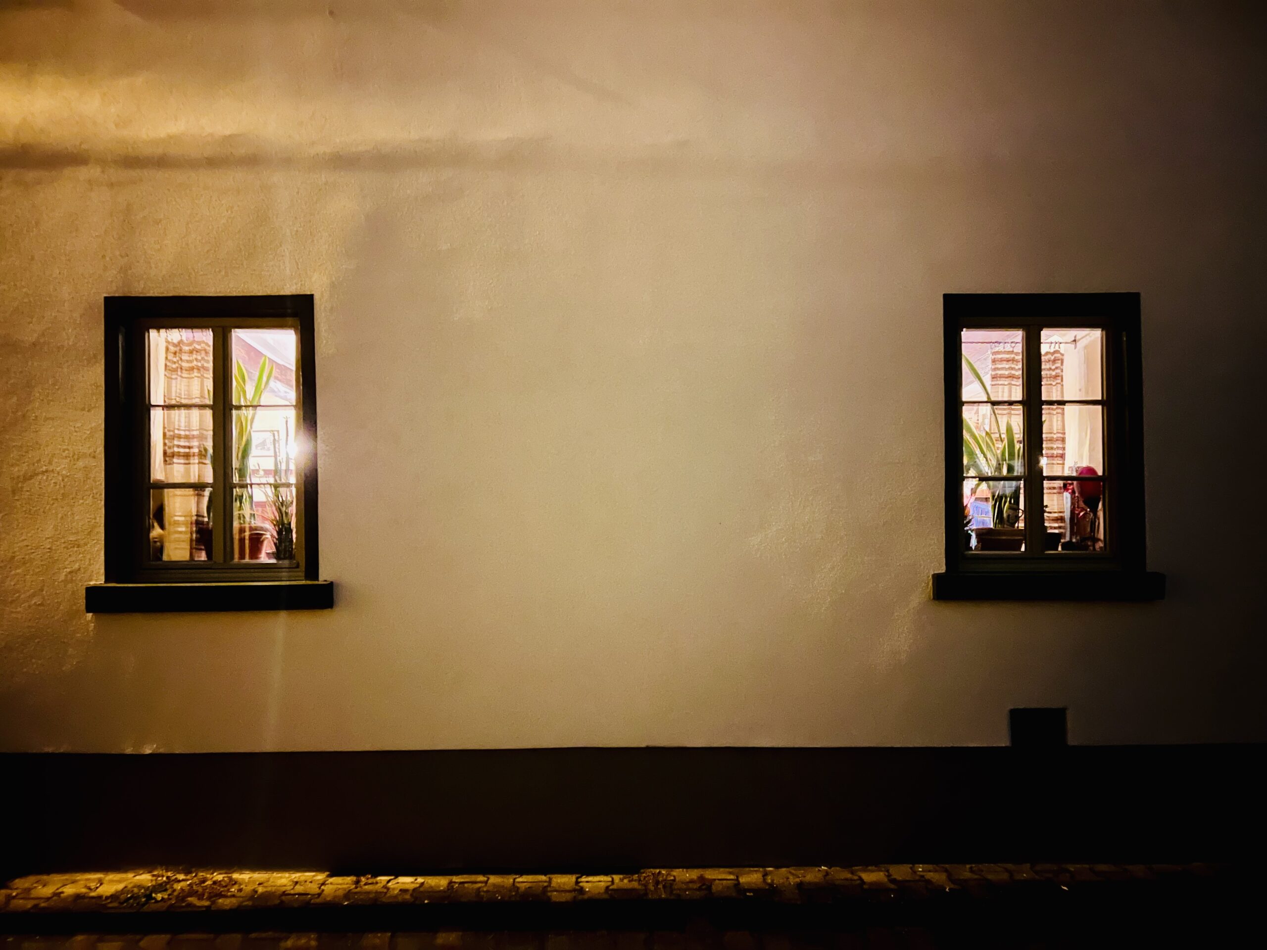 Zwei kleine Fenster in einer weißen Wand, durch die Licht aus der Gaststätte nach außen dringt. 